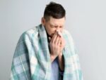 Man with plaid blanket suffering from runny nose from the flu on light background