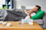 Man who is sick with the flu is lying on a couch while blowing his nose.
