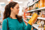 A young woman is reviewing the ingredient label of a food product at a grocery store.