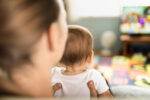 A parent holds young infant while they watch television screen.